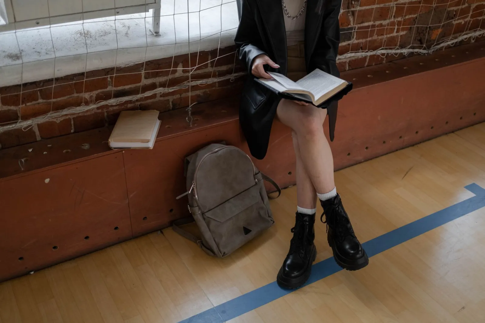 A student sitting on a bench reading a book with a backpack nearby, dressed in casual attire.